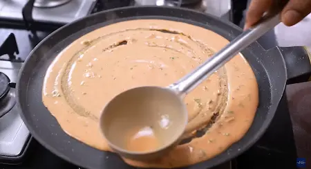 Pouring and spreading tomato dosa batter on a heated non-stick pan.