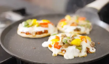 Cooking assembled mini pizzas on tawa with melted cheese.